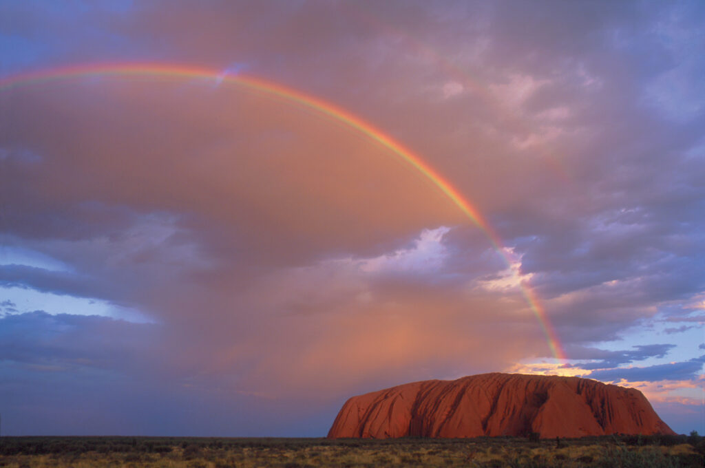 A landscape image of Uluru under a blue sky filled with dark peachy grey clouds. The arc of a rainbow is bursting out of the top of Uluru at the right of frame and ending on the left of frame.